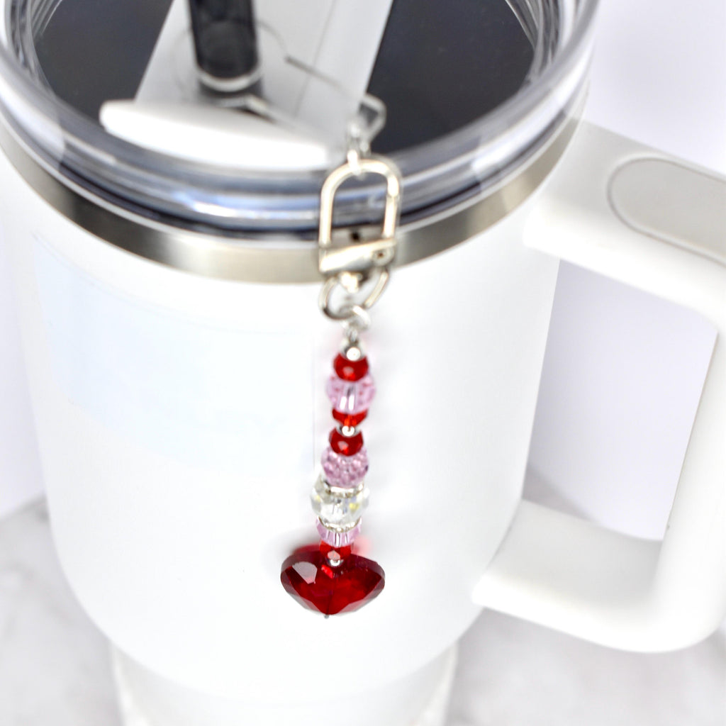 A white tumbler with a silver rim and handle, featuring a dangling charm. The charm is composed of red, pink, and clear beads, culminating in a large, heart-shaped red crystal. The charm is attached with a silver clasp.
