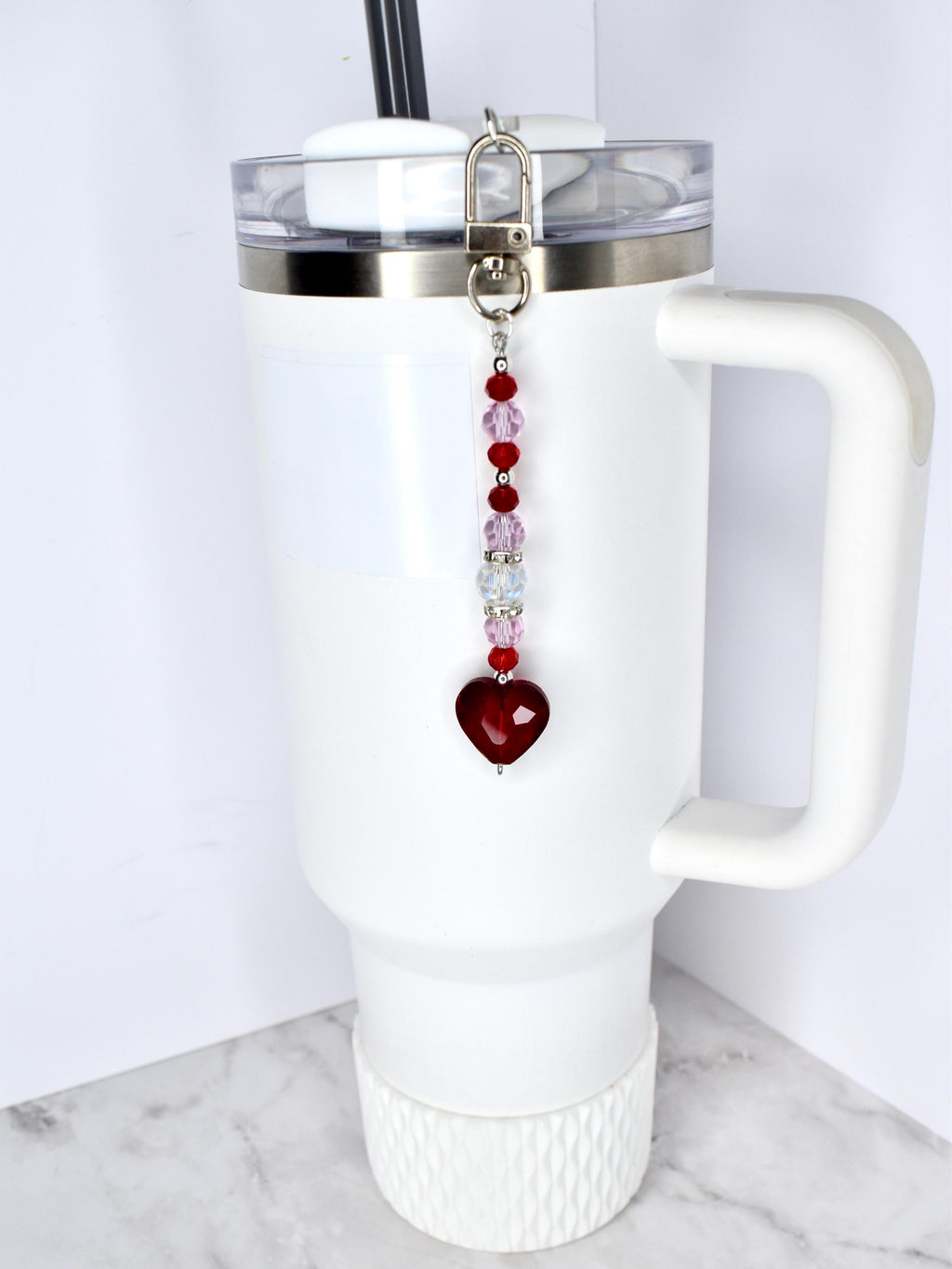 A white tumbler with a handle and a silver lid, featuring a beaded charm. The charm has a silver clasp, red and pink beads, and a heart-shaped pendant. The tumbler has a black straw.