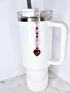A white tumbler with a handle and a silver lid, featuring a beaded charm. The charm has a silver clasp, red and pink beads, and a heart-shaped pendant. The tumbler has a black straw.