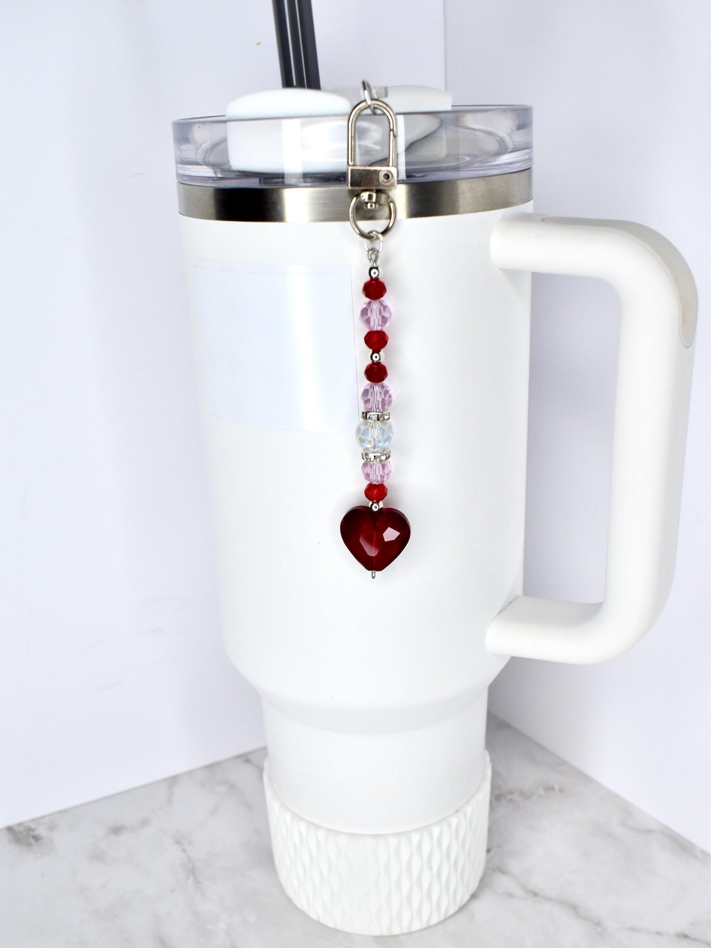 A white tumbler with a handle and a silver lid, featuring a beaded charm. The charm has a silver clasp, red and pink beads, and a heart-shaped pendant. The tumbler has a black straw.