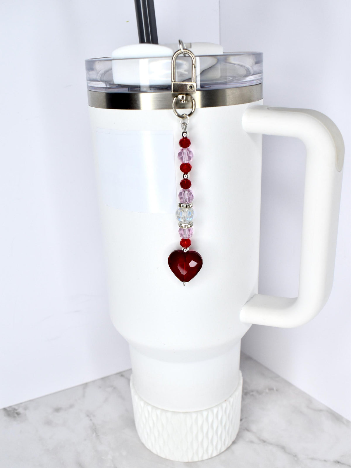 A white tumbler with a handle and a silver lid, featuring a beaded charm. The charm has a silver clasp, red and pink beads, and a heart-shaped pendant. The tumbler has a black straw.