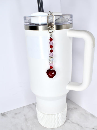 A white tumbler with a handle and a silver lid, featuring a beaded charm. The charm has a silver clasp, red and pink beads, and a heart-shaped pendant. The tumbler has a black straw.