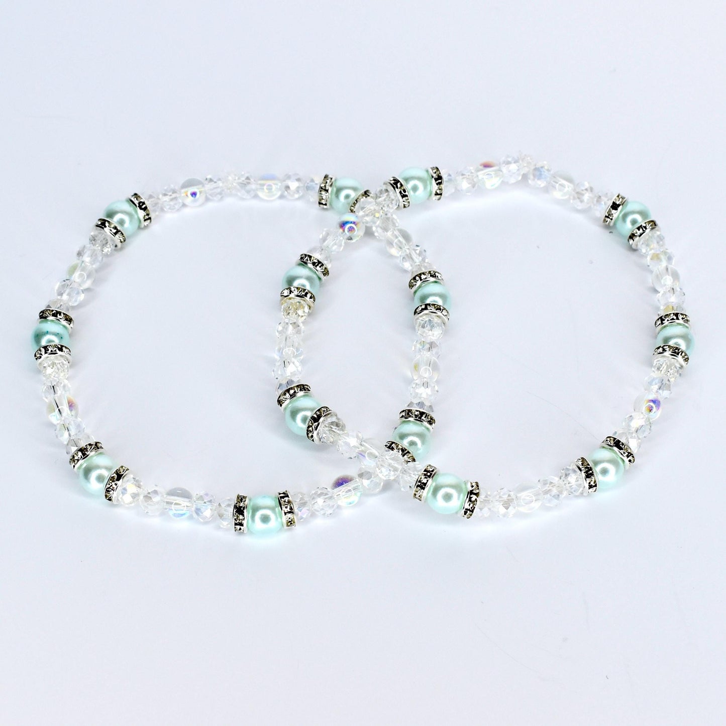 Two delicate bracelets with clear crystal beads and light blue pearl-like beads. The bracelets have silver-colored accents and are arranged in a figure-eight shape against a white background. The jewelry is likely for a special occasion.