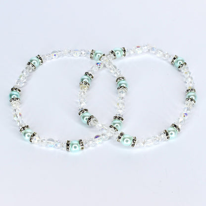 Two delicate bracelets with clear crystal beads and light blue pearl-like beads. The bracelets have silver-colored accents and are arranged in a figure-eight shape against a white background. The jewelry is likely for a special occasion.