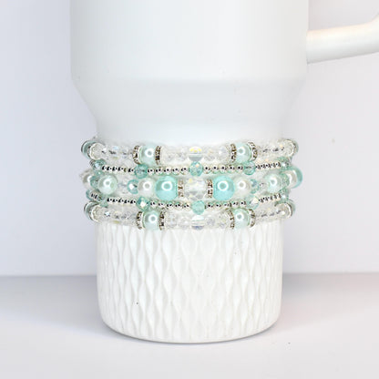 Four beaded bracelets in shades of white, clear, and light blue are stacked on a white Stanley tumbler. The bracelets feature clear beads, silver accents, and light blue pearl-like beads. The mug has a textured, diamond-like pattern.