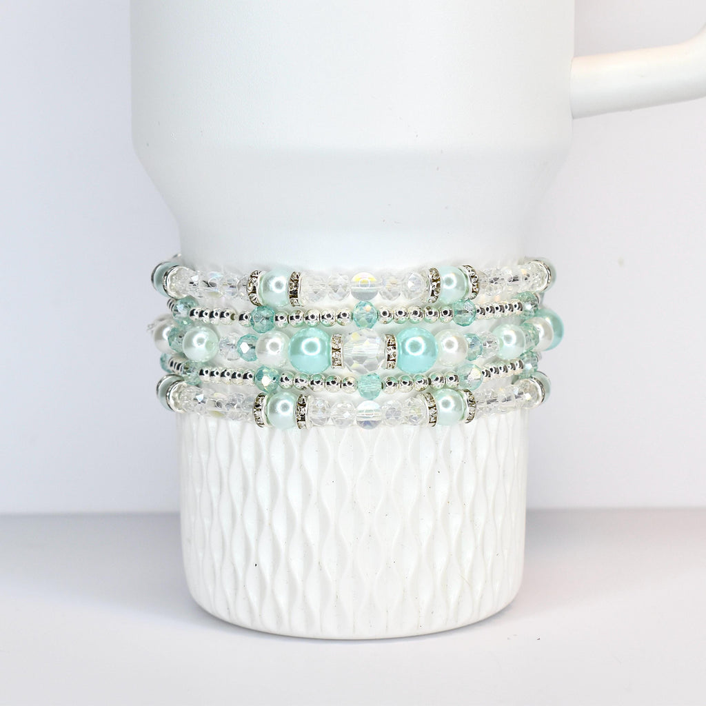 Four beaded bracelets in shades of white, clear, and light blue are stacked on a white Stanley tumbler. The bracelets feature clear beads, silver accents, and light blue pearl-like beads. The mug has a textured, diamond-like pattern.
