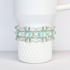 Four beaded bracelets in shades of white, clear, and light blue are stacked on a white Stanley tumbler. The bracelets feature clear beads, silver accents, and light blue pearl-like beads. The mug has a textured, diamond-like pattern.