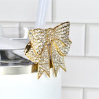 A gold-colored bow-shaped drink charm, encrusted with clear rhinestones, is attached to a white tumbler with a white straw. The charm adds a touch of elegance to the drinkware.