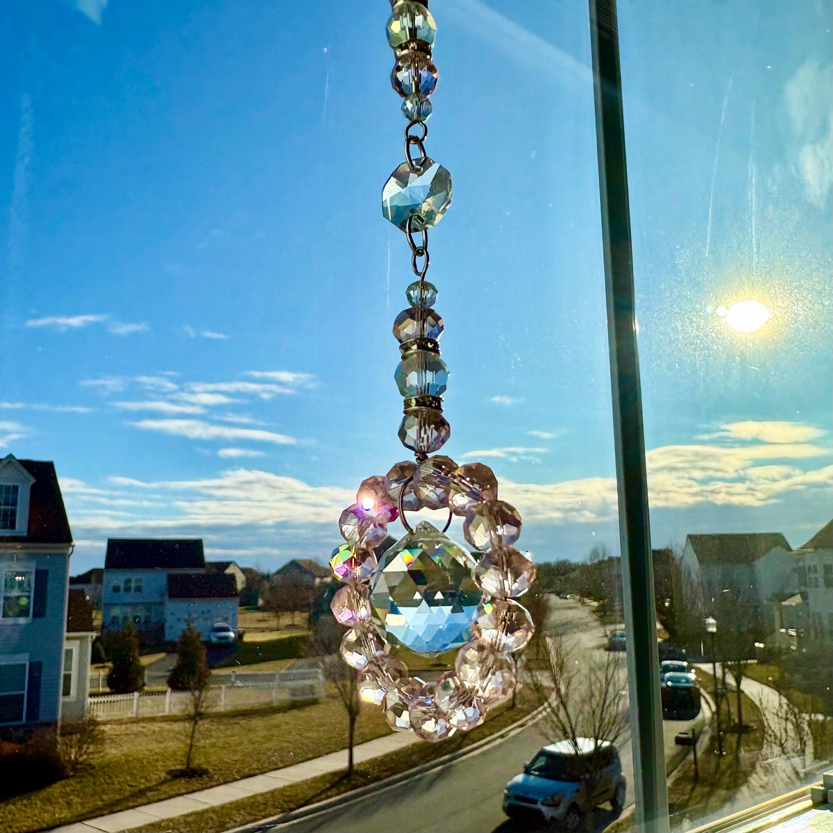 A decorative crystal suncatcher hanging in front of a window. The suncatcher features a large, faceted crystal ball surrounded by a ring of pink beads and smaller crystals. The sky and neighborhood are visible in the background.