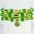A stack of beaded bracelets in shades of green and gold. The bracelets feature faceted beads, small gold beads, and gold heart and square accents. A green and gold shamrock charm hangs from the bottom bracelet.