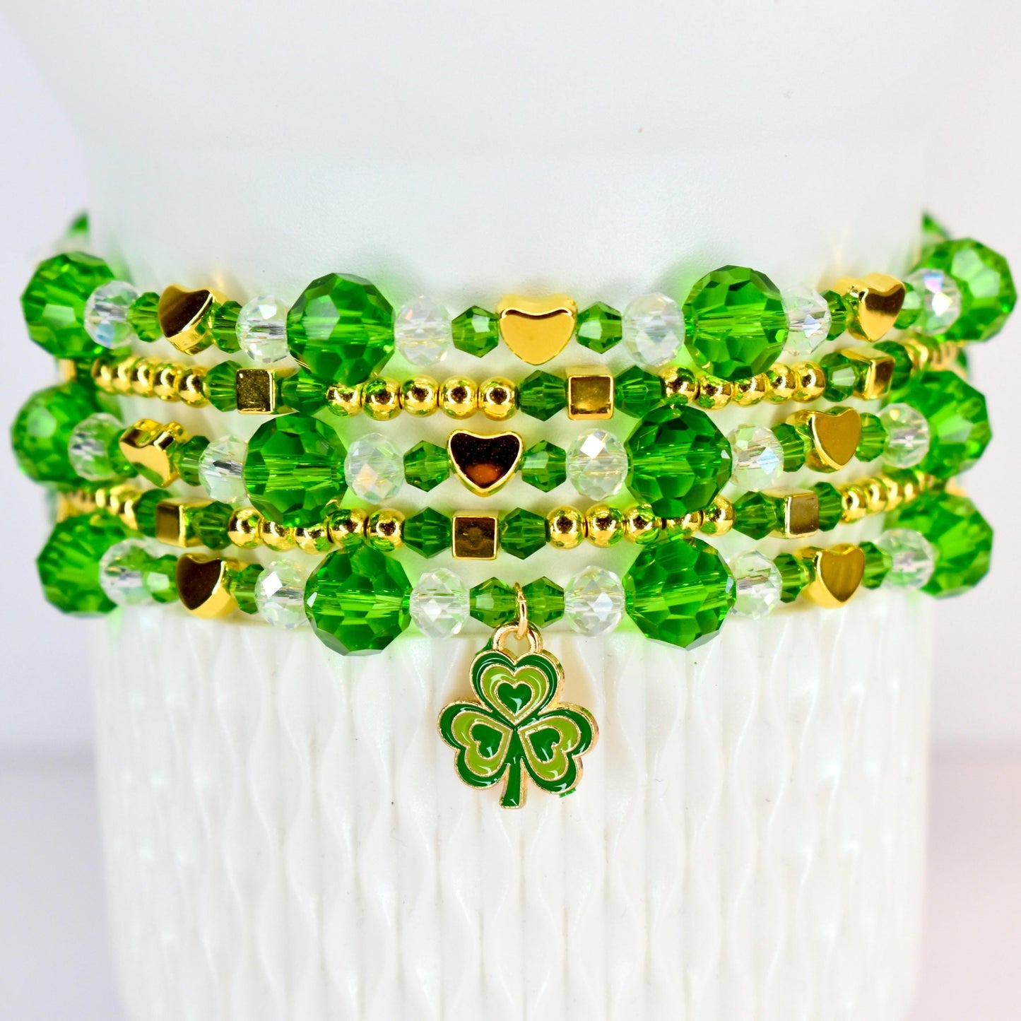 A stack of beaded bracelets in shades of green and gold. The bracelets feature faceted beads, small gold beads, and gold heart and square accents. A green and gold shamrock charm hangs from the bottom bracelet.