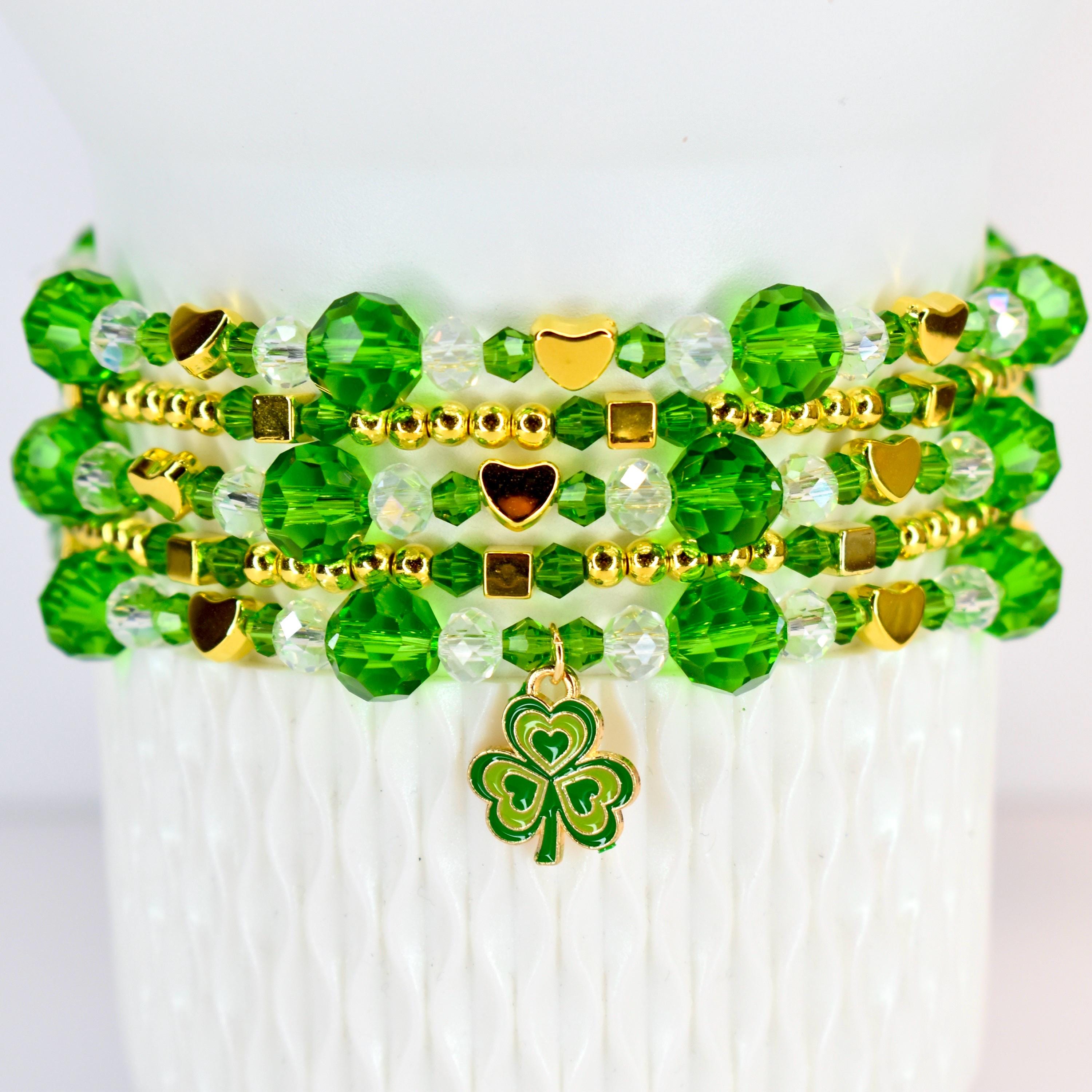 A stack of beaded bracelets in shades of green and gold. The bracelets feature faceted beads, small gold beads, and gold heart and square accents. A green and gold shamrock charm hangs from the bottom bracelet.