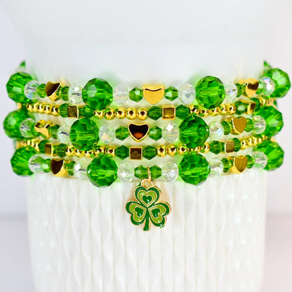 A stack of beaded bracelets in shades of green and gold. The bracelets feature faceted beads, small gold beads, and gold heart and square accents. A green and gold shamrock charm hangs from the bottom bracelet.