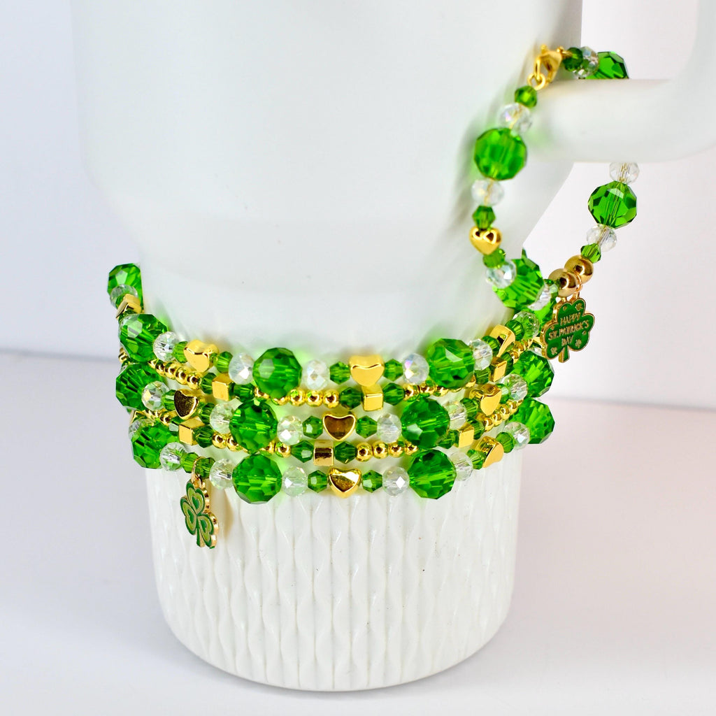 A stack of green beaded bracelets with gold accents and shamrock charms. The bracelets are draped over a white mug. The beads are various sizes and shapes, with gold heart-shaped charms.