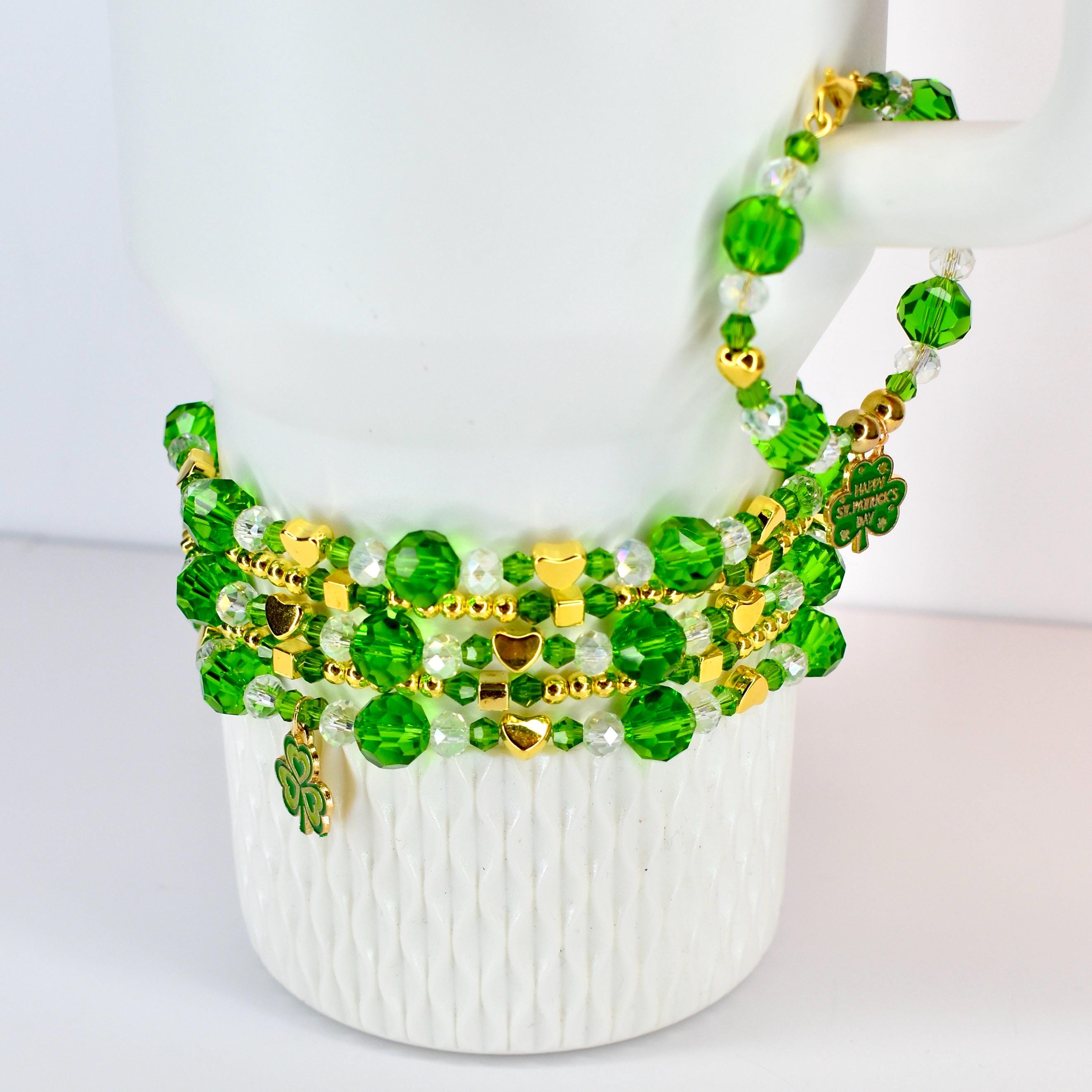 A stack of green beaded bracelets with gold accents and shamrock charms. The bracelets are draped over a white mug. The beads are various sizes and shapes, with gold heart-shaped charms.