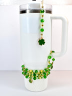 A white insulated tumbler adorned with green beaded accessories. A charm with a gold shamrock hangs from the lid. A beaded bracelet with green beads and gold accents wraps around the base, with a shamrock charm.