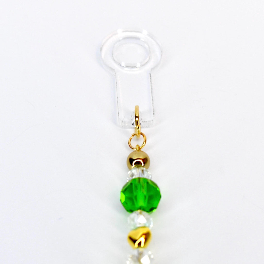 A clear, plastic charm with a gold-colored clasp and a beaded strand. The strand features a vibrant green faceted bead, gold-colored beads, and clear, iridescent beads. The charm is designed to be attached to a phone or other small device.