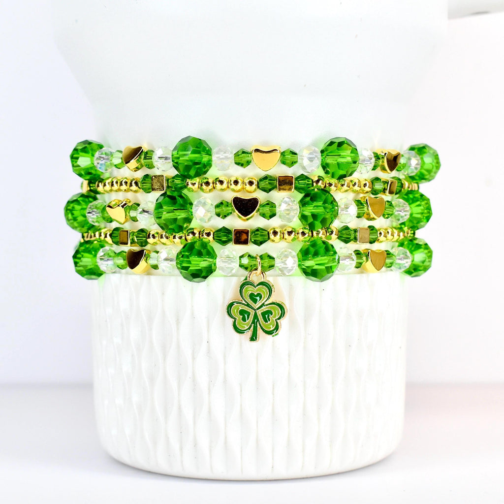 A stack of beaded bracelets in shades of green and clear, accented with gold-colored beads and charms. The bracelets feature round and faceted beads, with heart-shaped and square gold accents. A green and gold shamrock charm hangs from the bottom bracelet.