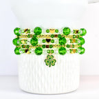 A stack of beaded bracelets in shades of green and clear, accented with gold-colored beads and charms. The bracelets feature round and faceted beads, with heart-shaped and square gold accents. A green and gold shamrock charm hangs from the bottom bracelet.