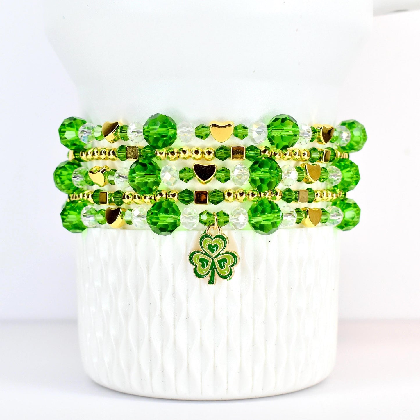 A stack of beaded bracelets in shades of green and clear, accented with gold-colored beads and charms. The bracelets feature round and faceted beads, with heart-shaped and square gold accents. A green and gold shamrock charm hangs from the bottom bracelet.