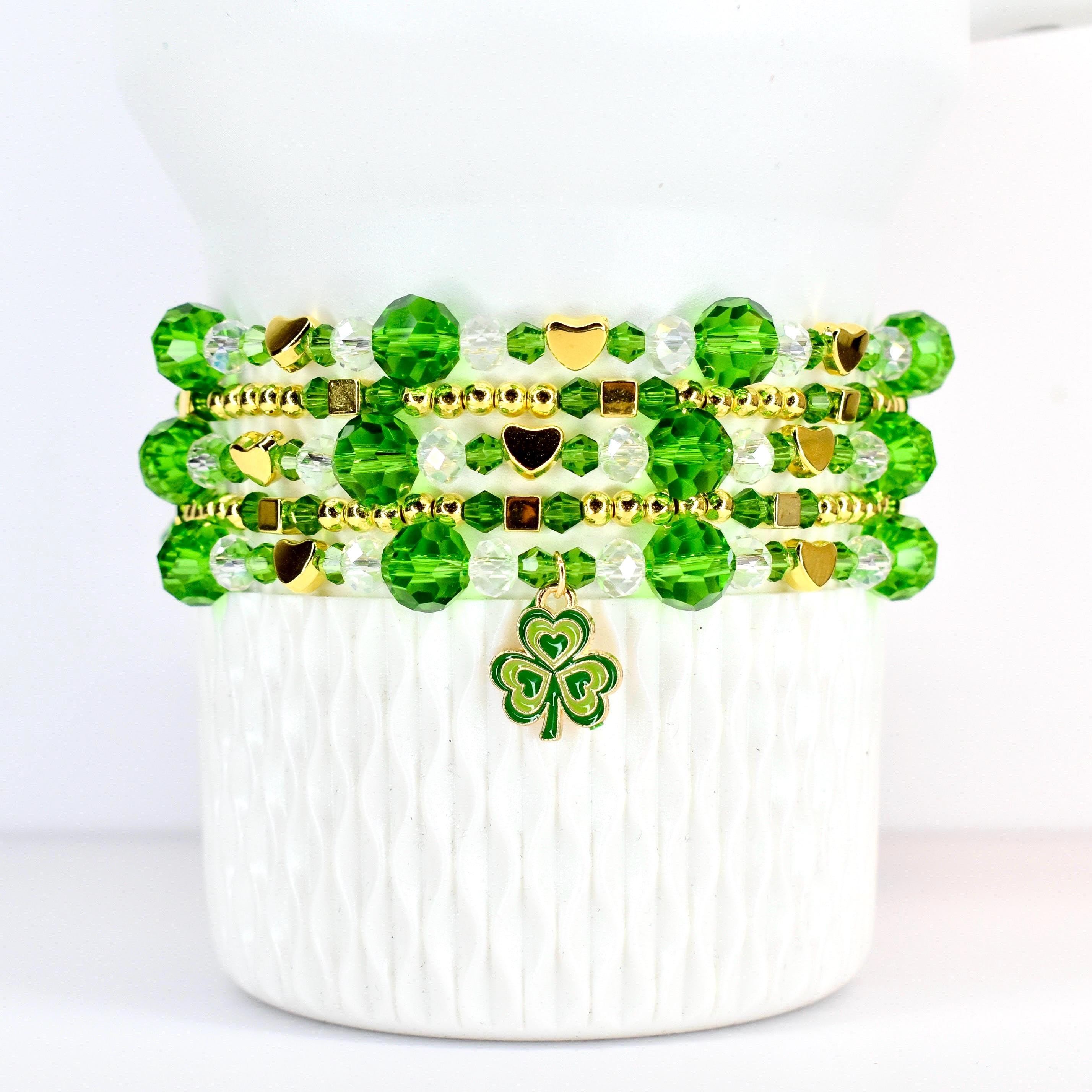 A stack of beaded bracelets in shades of green and clear, accented with gold-colored beads and charms. The bracelets feature round and faceted beads, with heart-shaped and square gold accents. A green and gold shamrock charm hangs from the bottom bracelet.