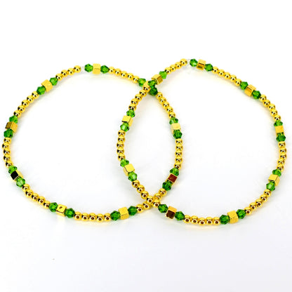Two gold-colored anklets with green and gold-colored beads. The anklets are made of small, round gold beads and square gold beads, interspersed with green, faceted beads. The anklets are displayed on a white background.