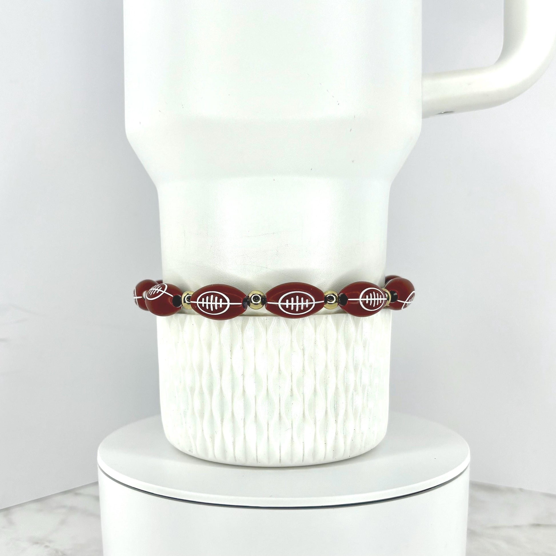 A football beaded bracelet featuring football-shaped beads with white detailing. The bracelet is strung with small gold-colored beads. The bracelet is displayed on a white ceramic mug.