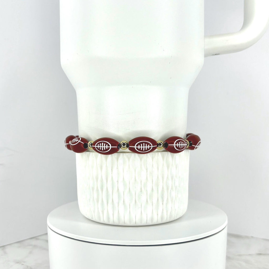 A football beaded bracelet featuring football-shaped beads with white detailing. The bracelet is strung with small gold-colored beads. The bracelet is displayed on a white ceramic mug.