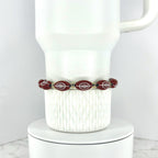 A football beaded bracelet featuring football-shaped beads with white detailing. The bracelet is strung with small gold-colored beads. The bracelet is displayed on a white ceramic mug.