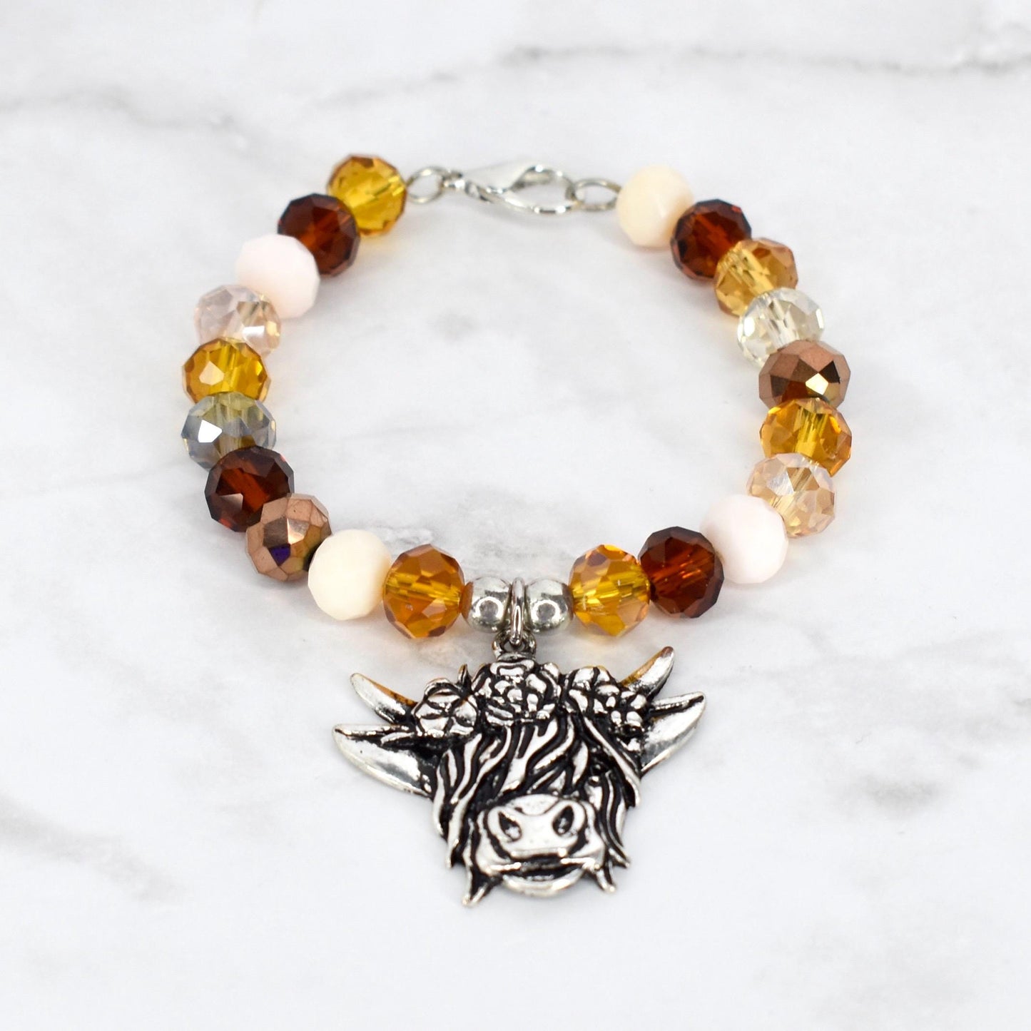 A beaded bracelet featuring a silver-toned charm of a Highland cow. The bracelet has a mix of amber, brown, cream, and clear crystal beads. The bracelet is secured with a silver-toned clasp.