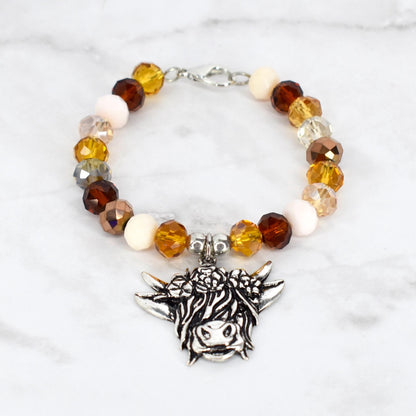 A beaded bracelet featuring a silver-toned charm of a Highland cow. The bracelet has a mix of amber, brown, cream, and clear crystal beads. The bracelet is secured with a silver-toned clasp.