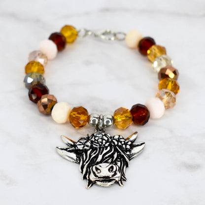 A beaded bracelet with a silver charm. The bracelet features a variety of colorful beads in shades of amber, brown, and cream. The charm is a detailed Highland cow head. The bracelet is on a white marble surface.