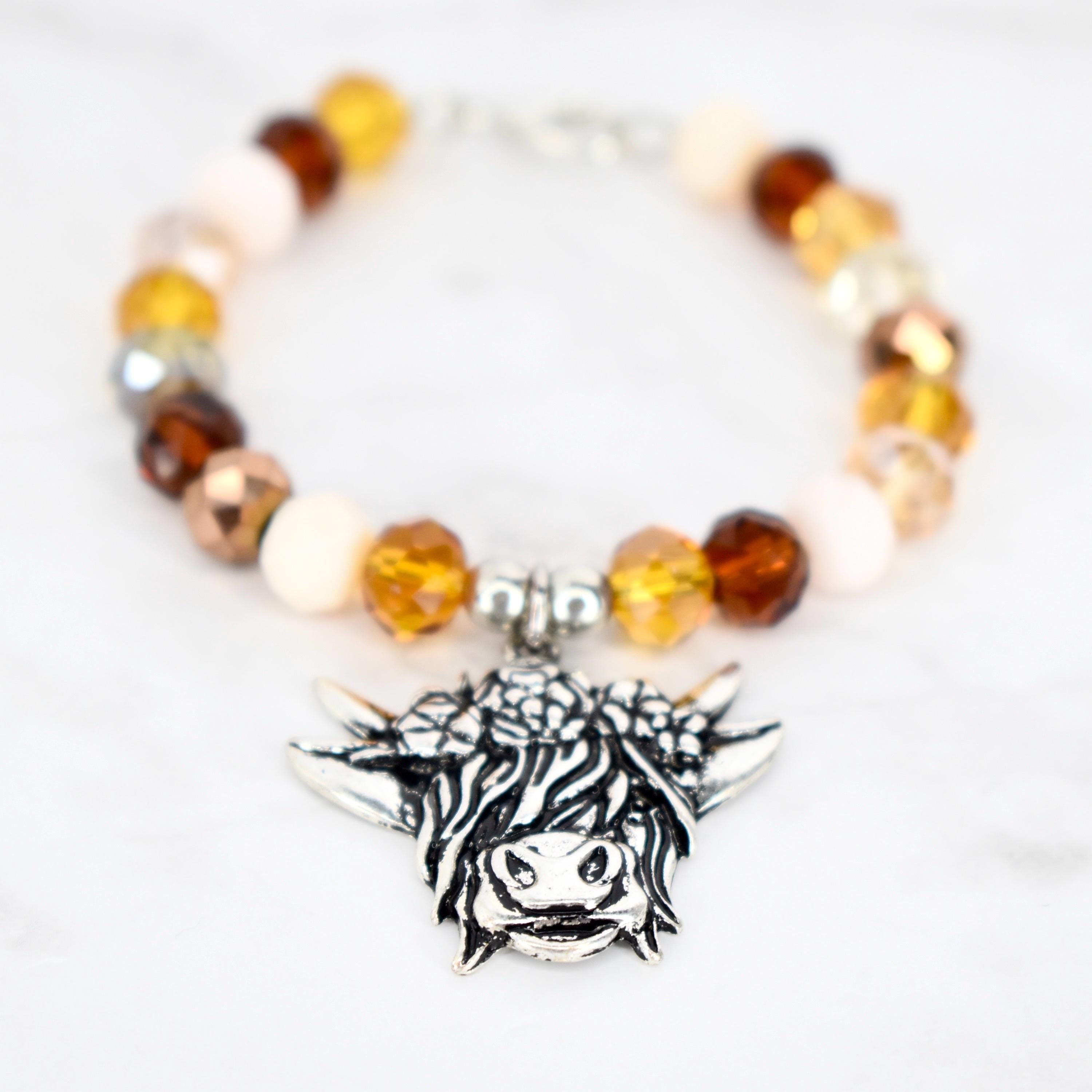 A beaded tumbler handle charm featuring a silver-toned Highland cow charm. The bracelet's beads are a mix of amber, brown, and cream colors. The charm is detailed with fur and facial features.