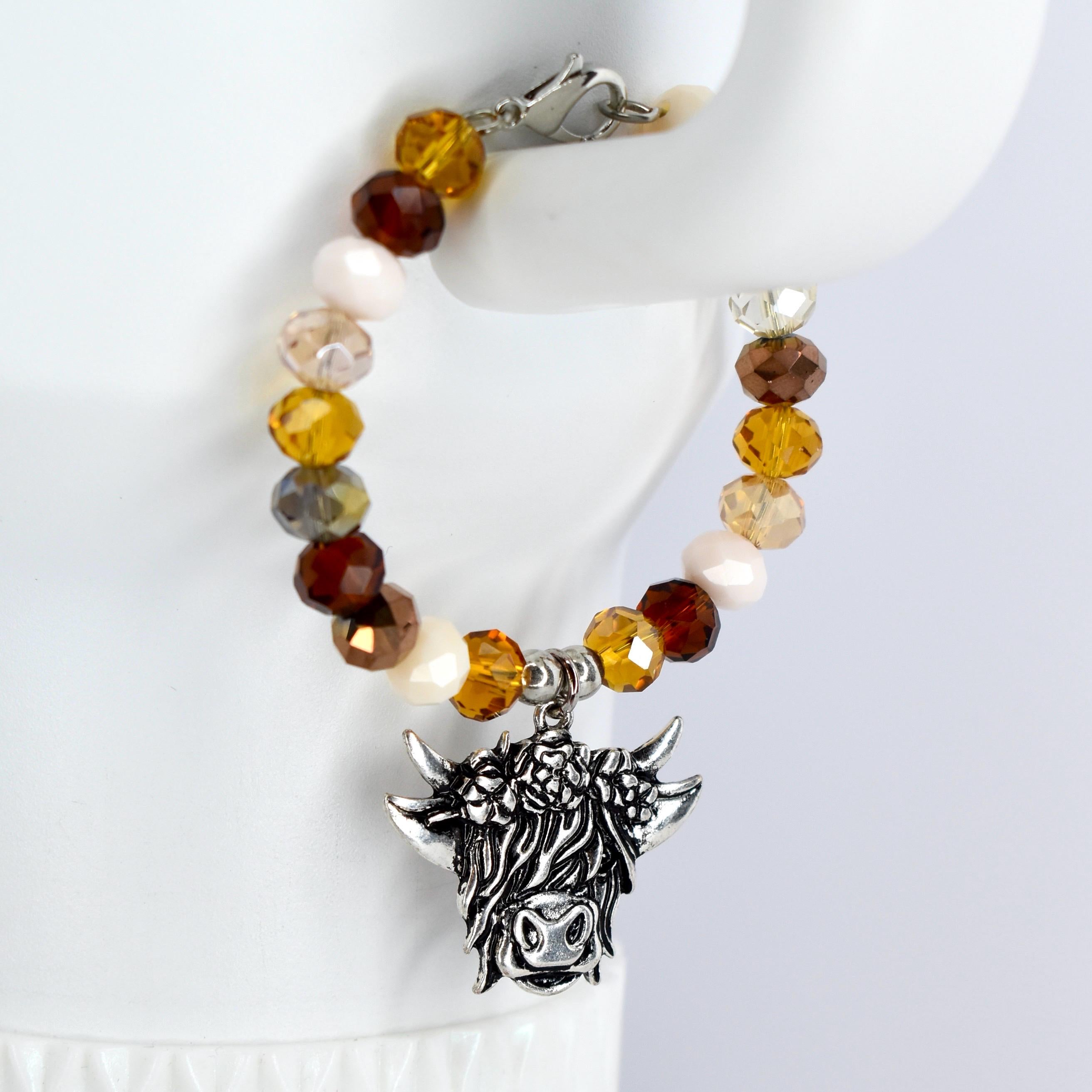A beaded bracelet with a silver-colored charm. The bracelet features a variety of colorful beads in shades of brown and cream. The charm is a detailed depiction of a Highland cow's head.