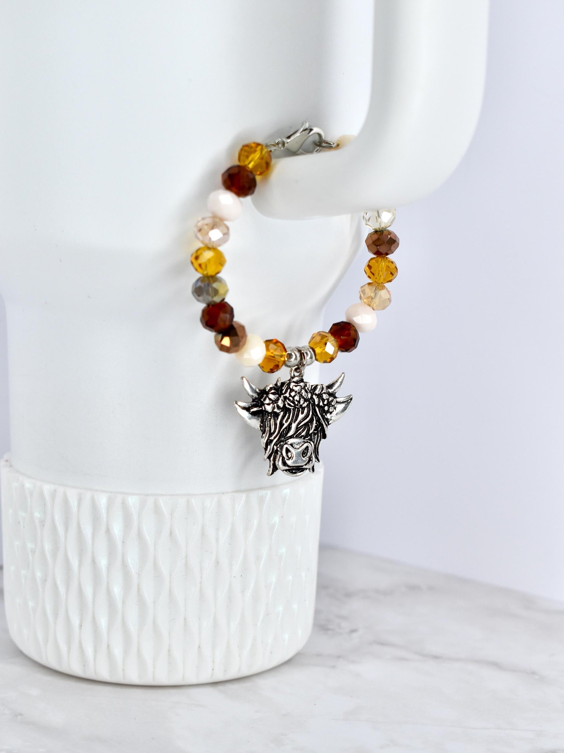 A beaded bracelet with a silver-colored charm of a Highland cow. The bracelet features a mix of amber, brown, cream, and clear beads. The charm is detailed with the cow's face and horns, and the bracelet is secured with a silver clasp.