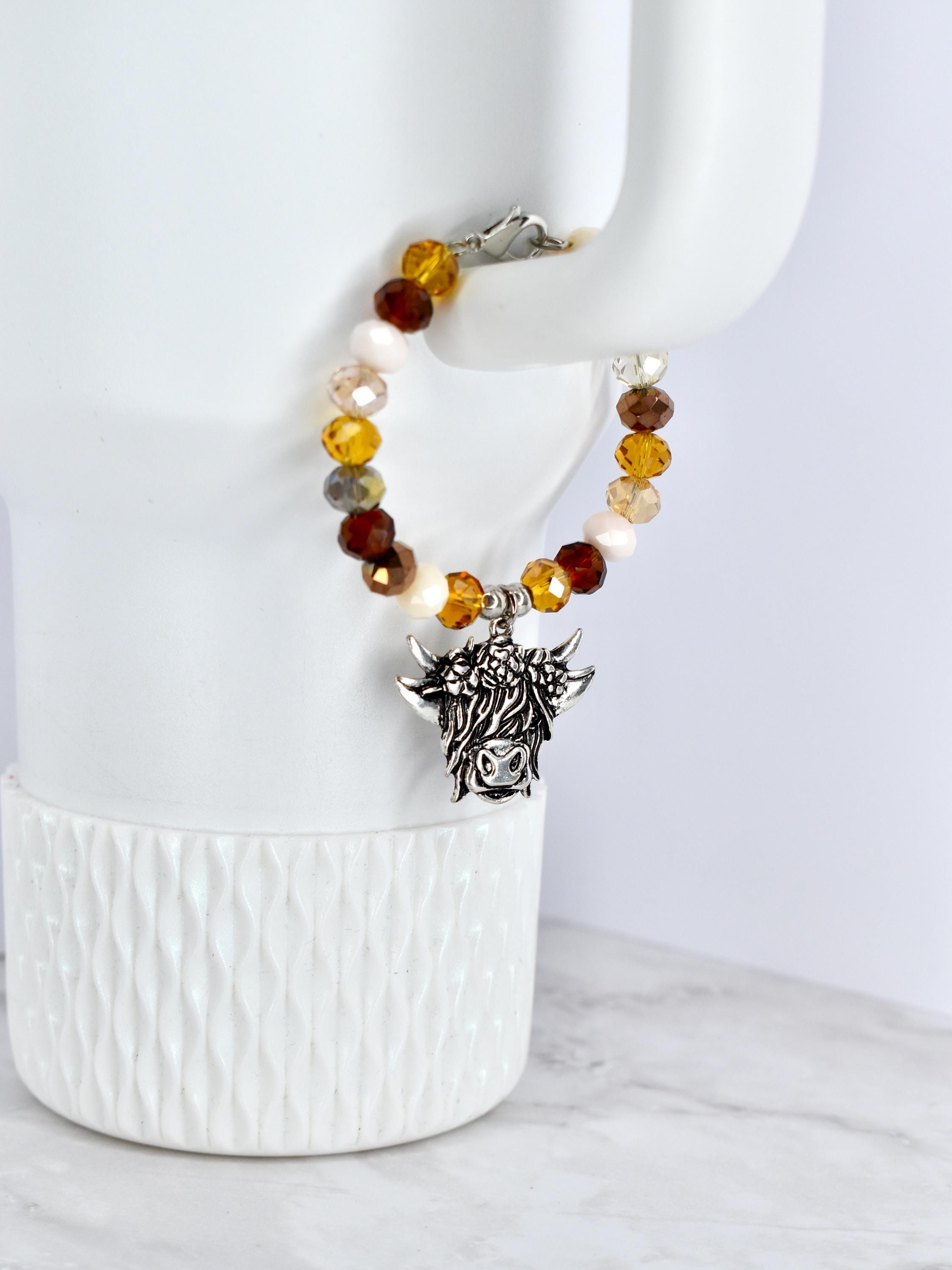 A beaded bracelet with a silver-colored charm of a Highland cow. The bracelet features a mix of amber, brown, cream, and clear beads. The charm is detailed with the cow's face and horns, and the bracelet is secured with a silver clasp.