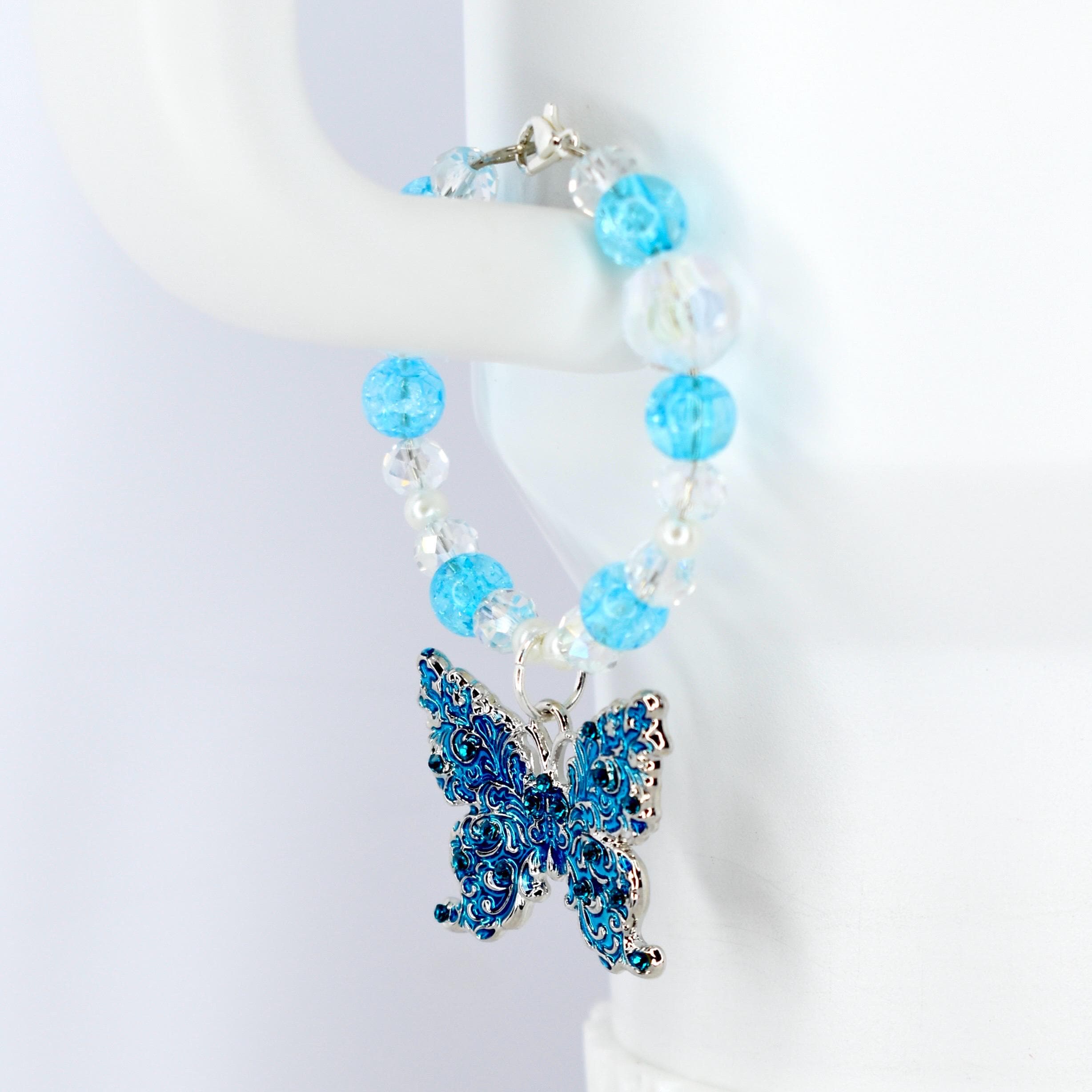 A beaded tumbler handle bracelet with a blue butterfly charm. The bracelet features light blue and clear beads. The butterfly charm is blue with silver accents and intricate detailing. The bracelet is displayed on a white surface.