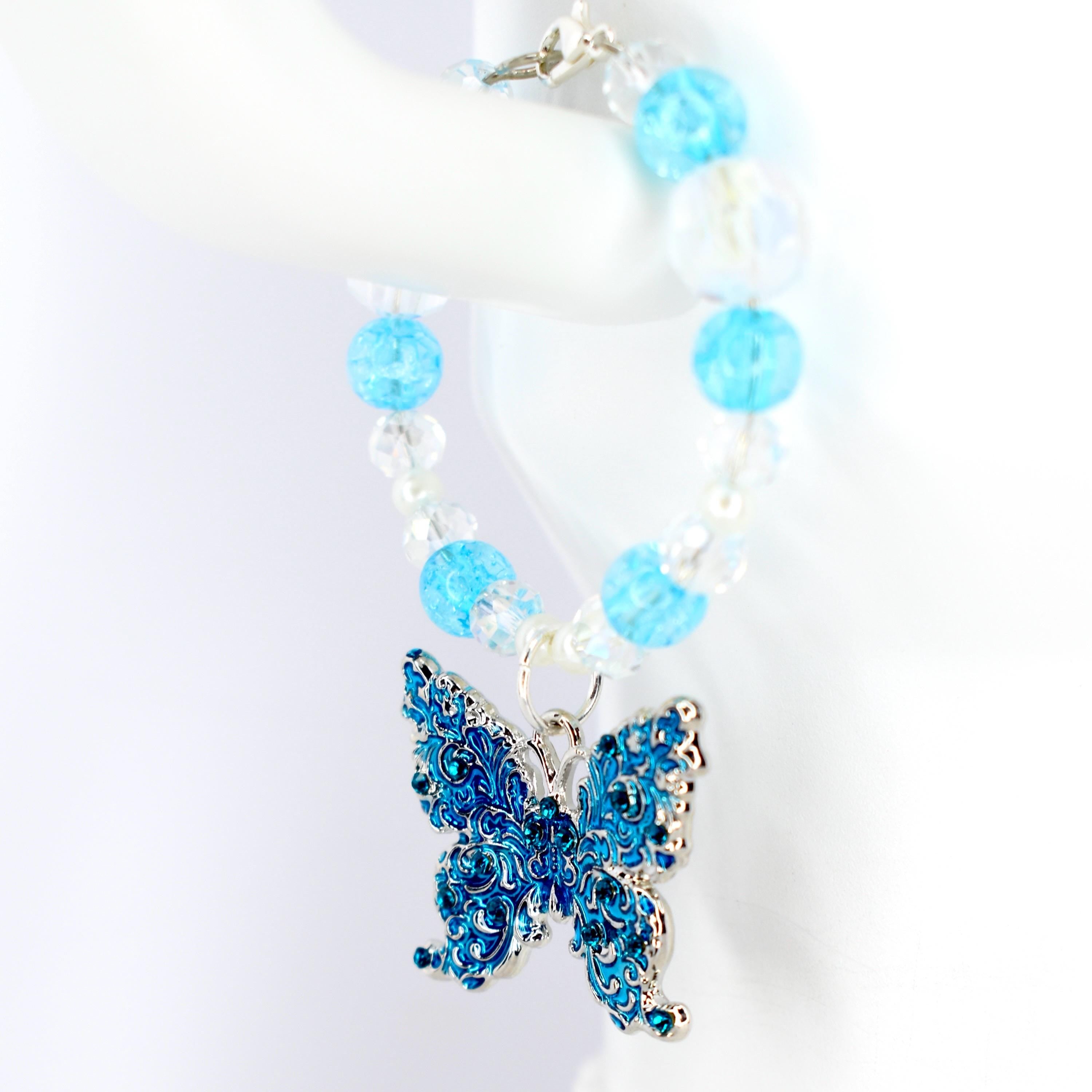 A blue and silver butterfly charm tumbler handle bracelet. The tumbler bracelet features a detailed, ornate butterfly pendant with blue accents and clear and blue beaded accents. The bracelet is held up against a white background.