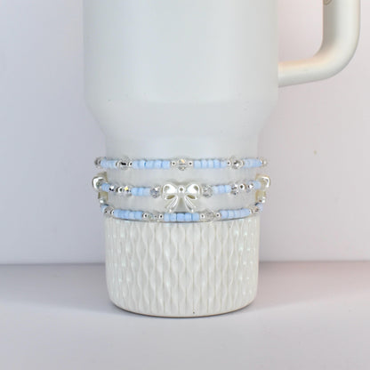 A white tumbler with three beaded bands. The bands feature light blue beads, silver accents, and a silver bow. The tumbler has a textured pattern on the bottom half.