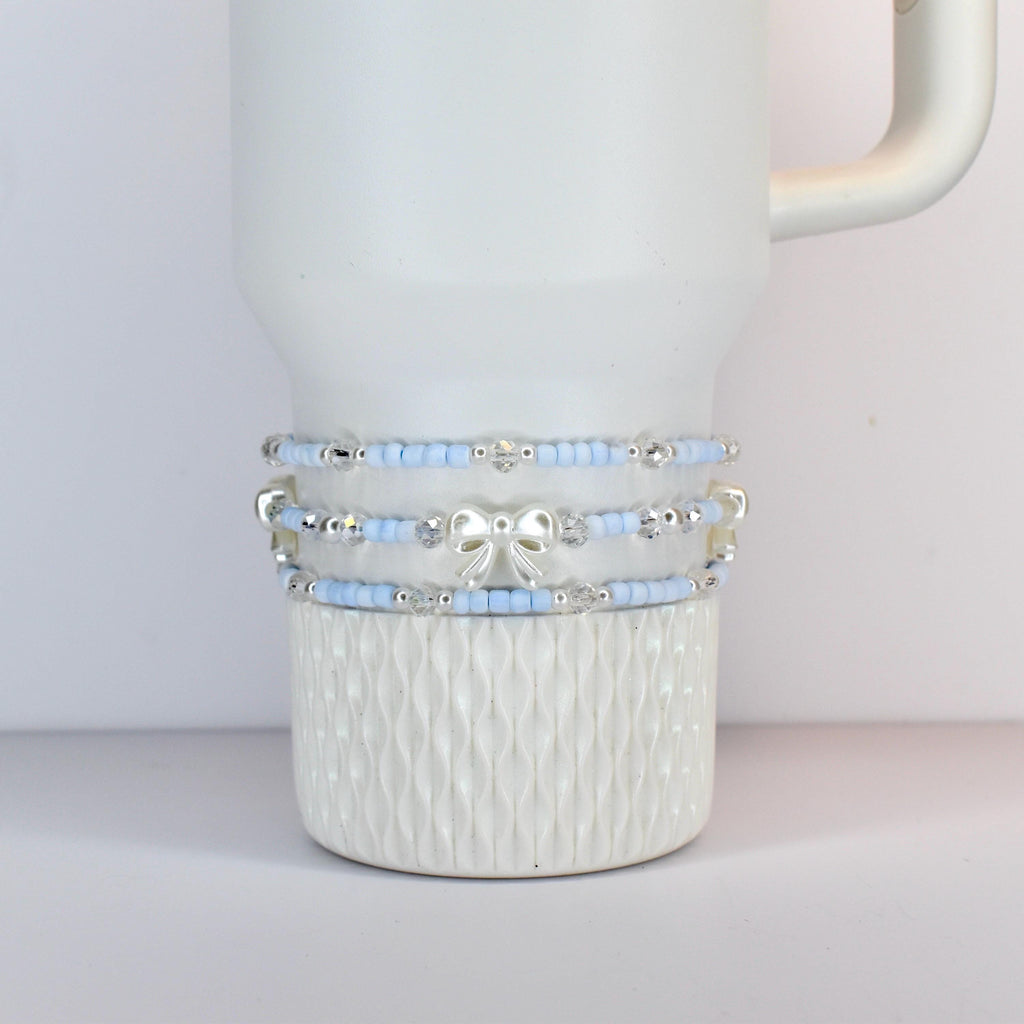 A white tumbler with three beaded bands. The bands feature light blue beads, silver accents, and a silver bow. The tumbler has a textured pattern on the bottom half.