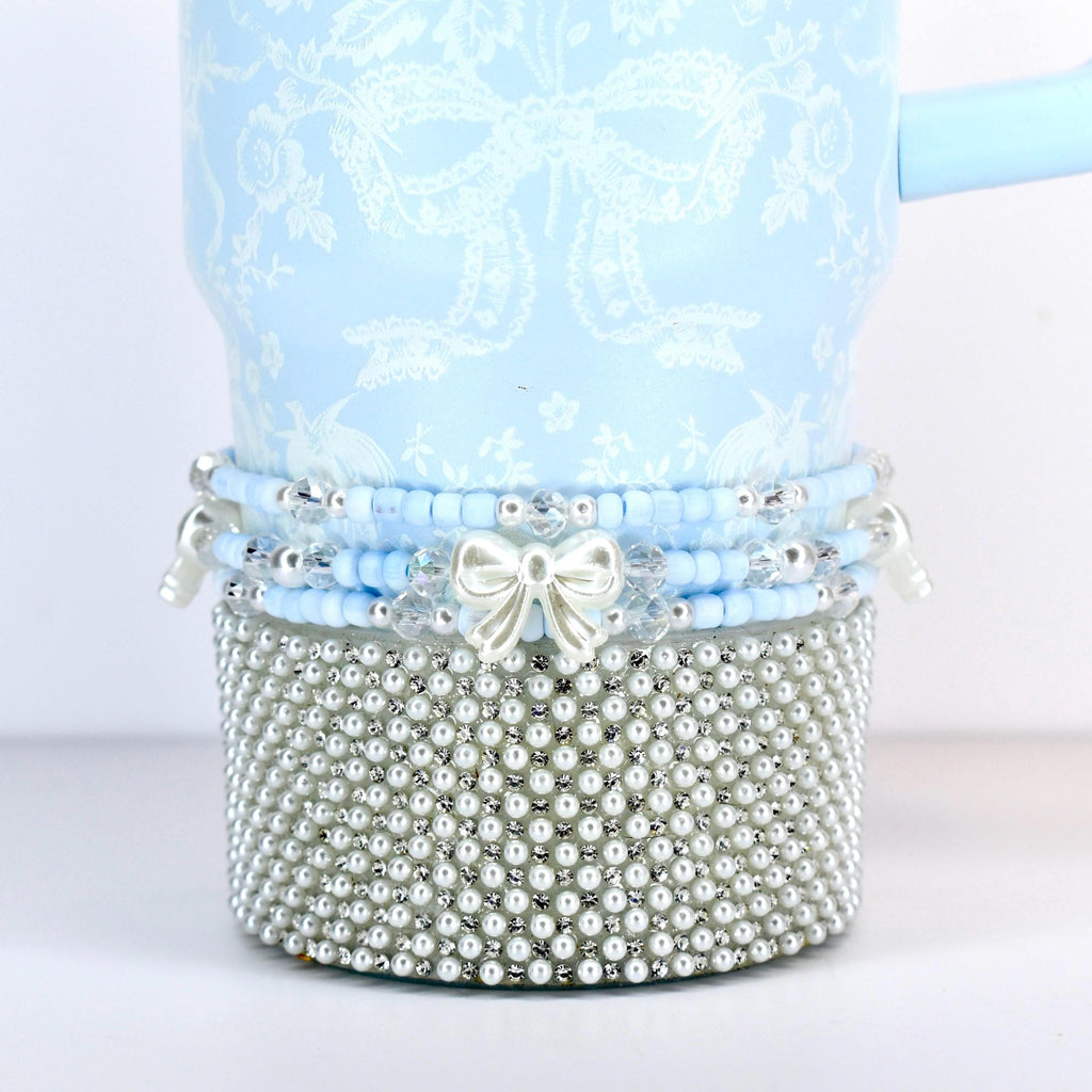 A light blue tumbler with a white floral and bow pattern. The bottom is covered in small, silver-colored faux pearls. A band of blue beads and a white bow accent the tumbler.