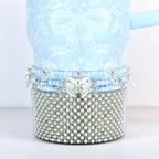 A light blue tumbler with a white floral and bow pattern. The bottom is covered in small, silver-colored faux pearls. A band of blue beads and a white bow accent the tumbler.