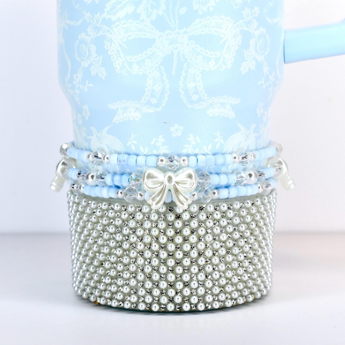 A light blue tumbler with a white floral and bow pattern. The bottom is covered in small, silver-colored faux pearls. A band of blue beads and a white bow accent the tumbler.