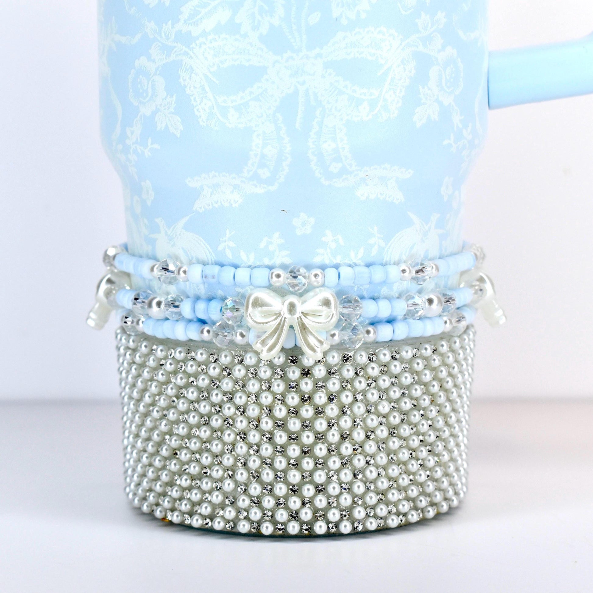 A light blue tumbler with a white floral and bow pattern. The bottom is covered in small, silver-colored faux pearls. A band of blue beads and a white bow accent the tumbler.