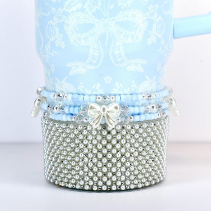 A light blue tumbler with a white floral and bow pattern. The bottom is covered in small, silver-colored faux pearls. A band of blue beads and a white bow accent the tumbler.