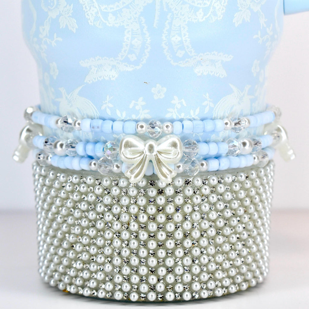 A light blue decorative item with a white floral pattern. The item is adorned with layers of blue beads, clear crystals, and a silver bow. The base is covered in small, white pearls, creating a luxurious aesthetic.