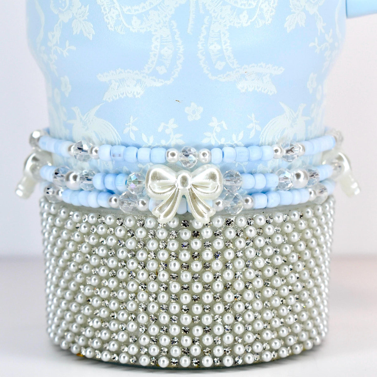 A light blue decorative item with a white floral pattern. The item is adorned with layers of blue beads, clear crystals, and a silver bow. The base is covered in small, white pearls, creating a luxurious aesthetic.