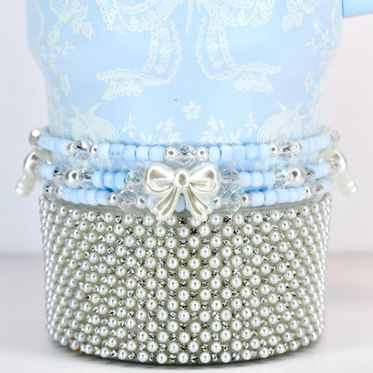 A light blue decorative item with a white floral pattern. The item is adorned with layers of blue beads, clear crystals, and a silver bow. The base is covered in small, white pearls, creating a luxurious aesthetic.