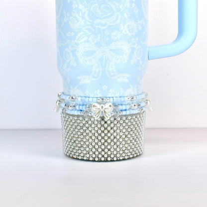 A light blue tumbler with a floral and bow pattern. The bottom section is covered in silver-colored faux pearls. A light blue handle is attached to the side. The bottom of the tumbler has a beaded design with a silver bow.