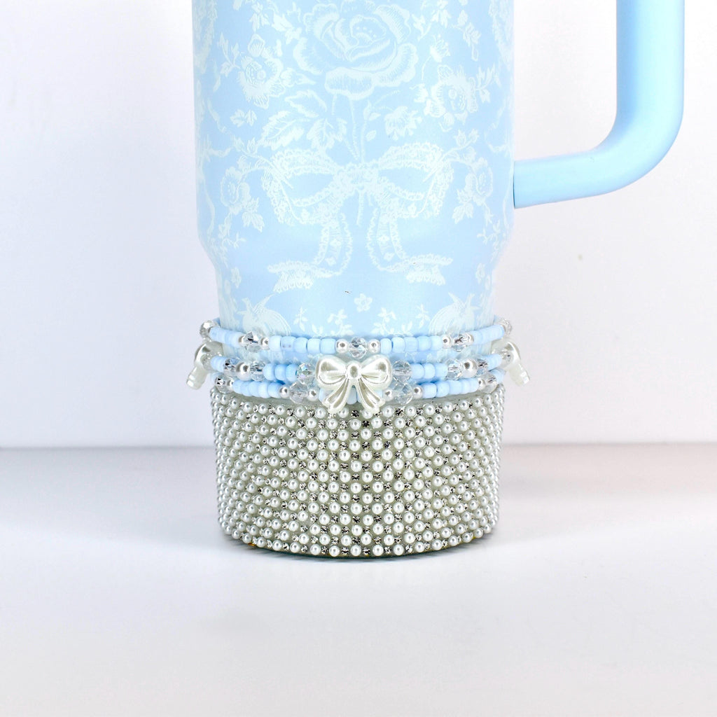 A light blue tumbler with a floral and bow pattern. The bottom section is covered in silver-colored faux pearls. A light blue handle is attached to the side. The bottom of the tumbler has a beaded design with a silver bow.